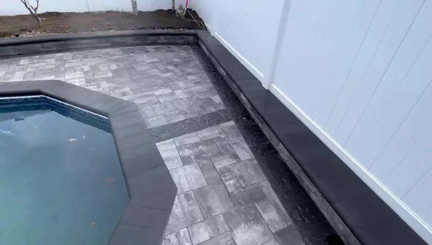 Octagonal pool with black coping and a paver deck against a white wall.