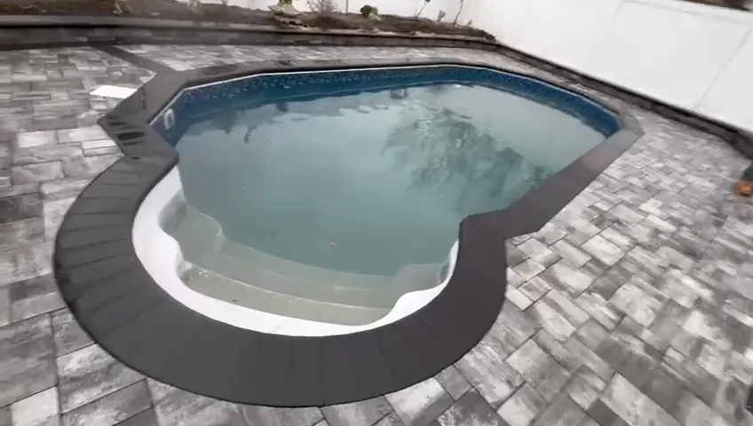 Paver pool surround with black coping, with the lawn and fence beyond.