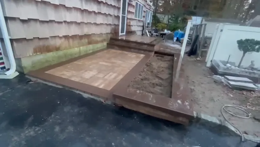 Raised paver platform going in against the house, with soil beds and site materials around it.